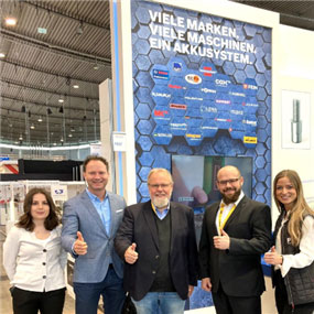 Five people pose in front of a wall display at a trade fair.