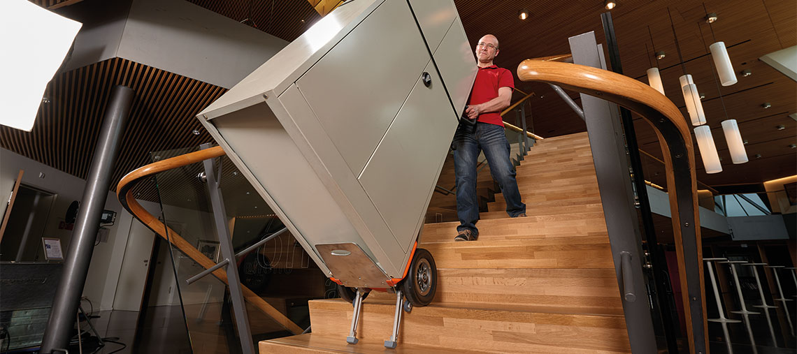 Mann trägt einen silbernen Schrank eine Holztreppe hoch.