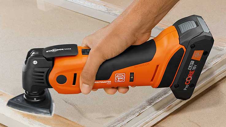 Eine Hand hält einen orangefarbenen Bosch Akku-Multitool auf Holz.