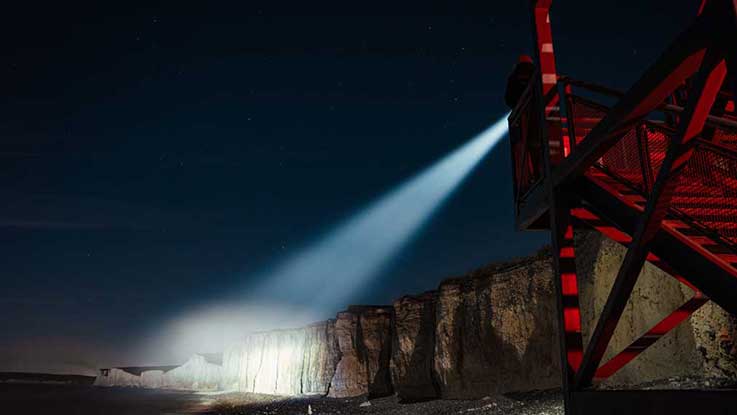 Ein roter Leuchtturm strahlt Licht auf Klippen und Strand bei Nacht.