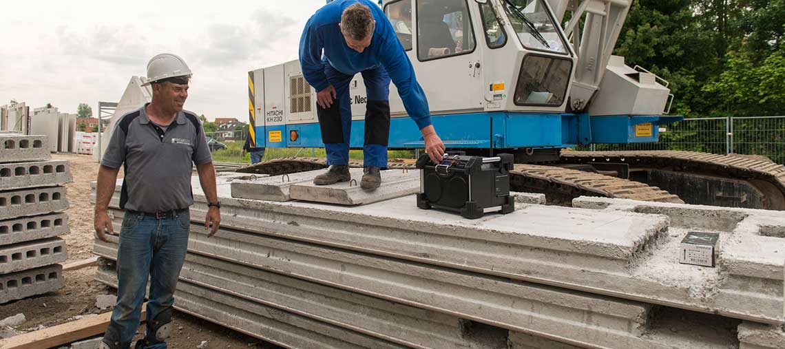 Zwei Bauarbeiter stehen neben Betonplatten und einem Kran.