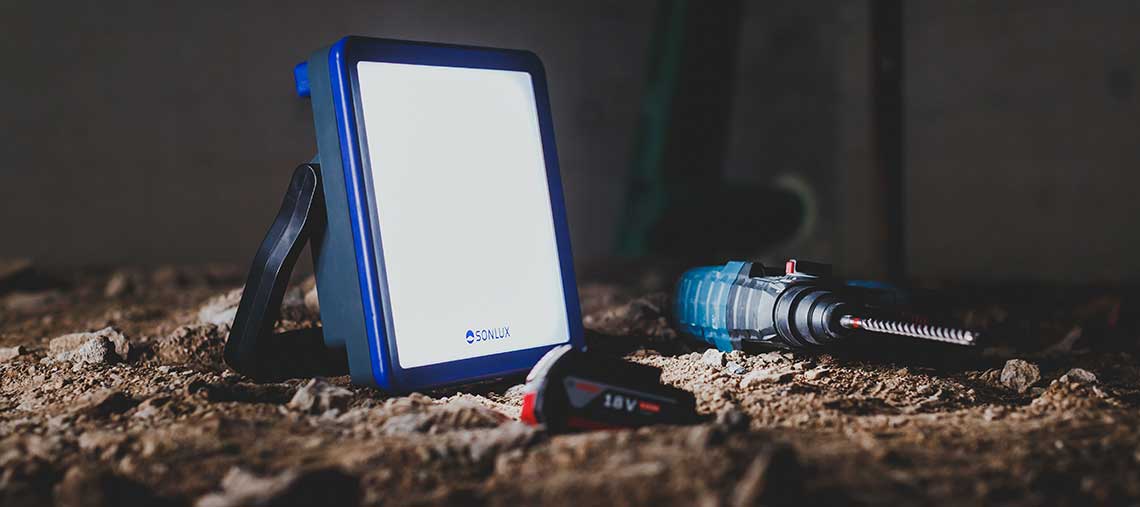 Eine blaue SONLUX Baustellenlampe steht auf dem Boden neben einem Akku-Bohrer.