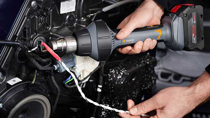 Ein grauer Steinel Heißluftfön wird an einem Kabelanschluss gehalten.